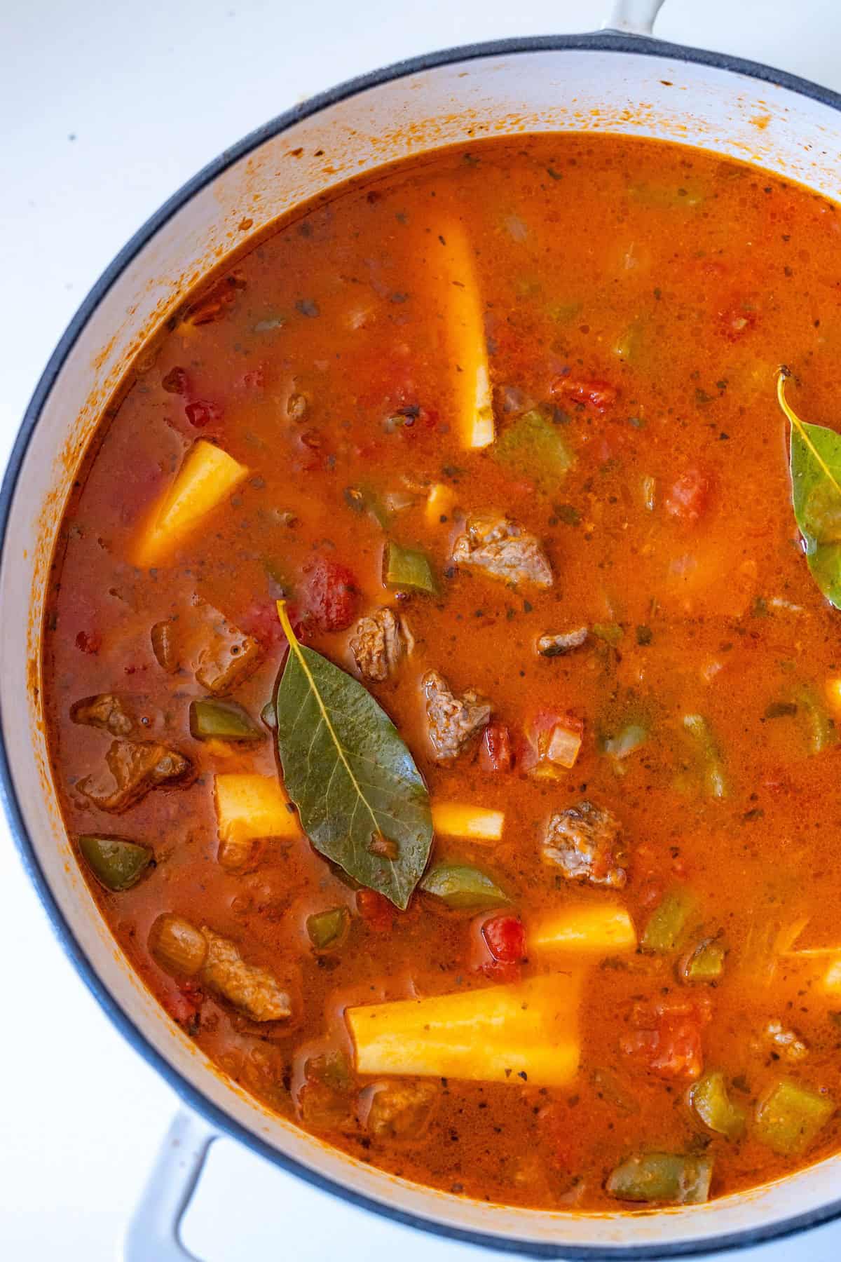 A pot of beef stew brimming with chunks of tender meat, yellow potatoes, tomatoes, green peppers, and bay leaves in a rich, spiced broth.