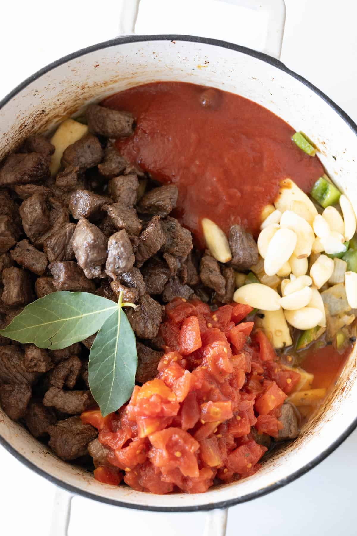 Pot with beef stew, featuring tender cooked beef, green peppers, sliced garlic, diced tomatoes in a rich tomato sauce, enhanced by a bay leaf.