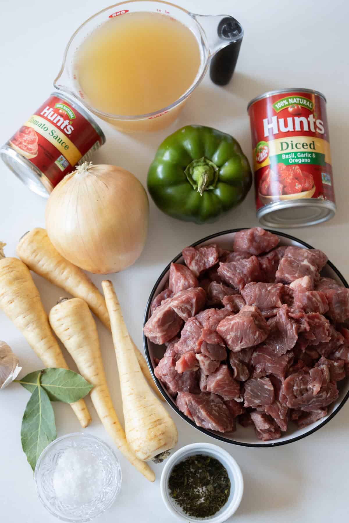 A vibrant medley of stew ingredients awaits, featuring succulent raw beef cubes, aromatic onion, crisp green bell pepper, and sweet parsnips. Canned tomatoes mingle with rich beef broth and fragrant bay leaves, all poised on a light surface to create a harmonious blend of flavors.