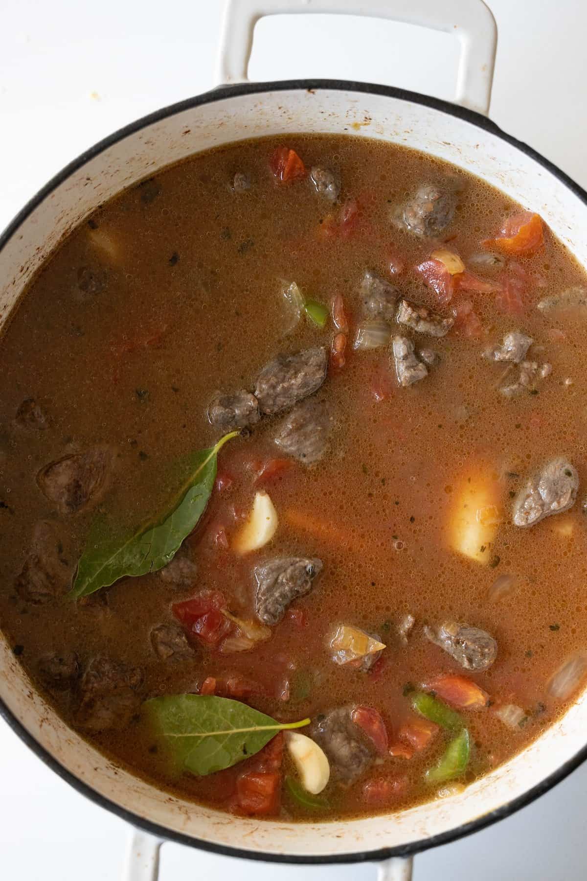 A pot of beef stew containing chunks of meat, sliced tomatoes, garlic cloves, a bay leaf, and visible spices in a savory broth.