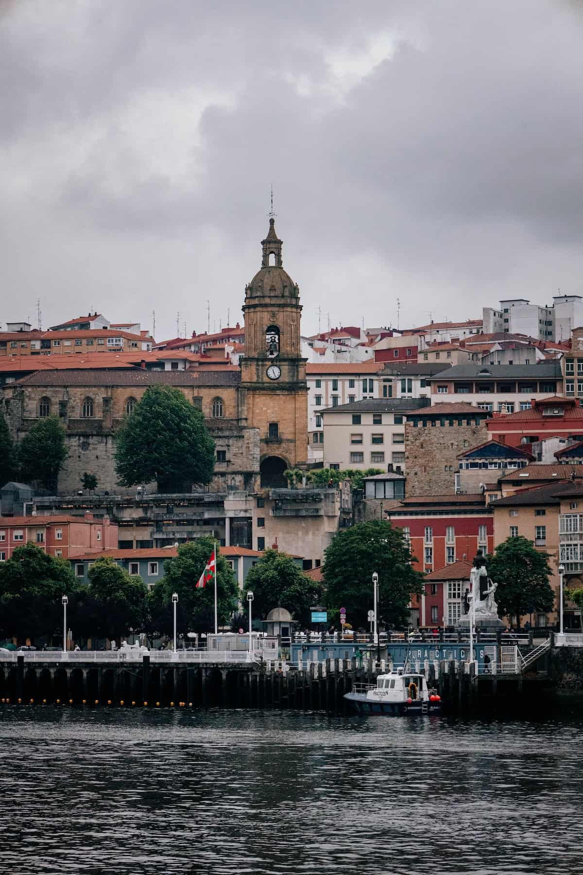 A waterfront town with a clock tower, multiple buildings, trees, and a docked boat on a cloudy day.