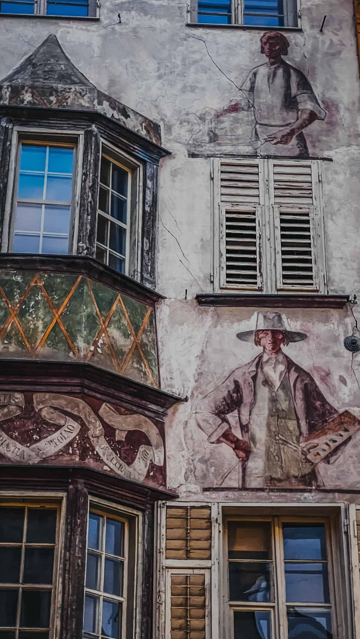 The facade of an old building in Bolzano features murals depicting human figures, decorative patterns, and multiple windows with shutters, reminiscent of a Christmas market's charm.