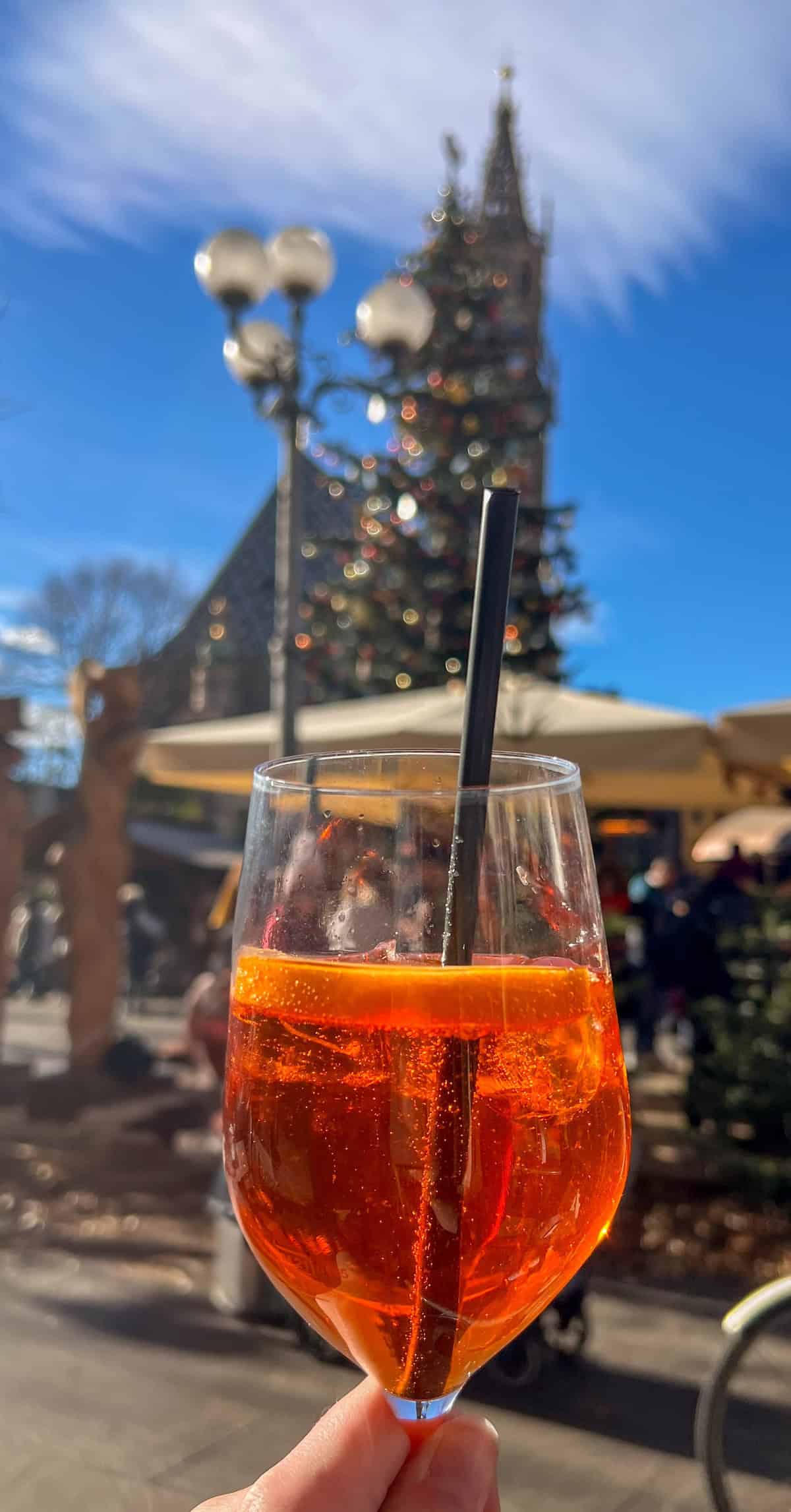 A glass of orange cocktail with a straw is held up in an outdoor setting, with a blurred view of a Christmas tree and the charming buildings of Bolzano's market in the background.