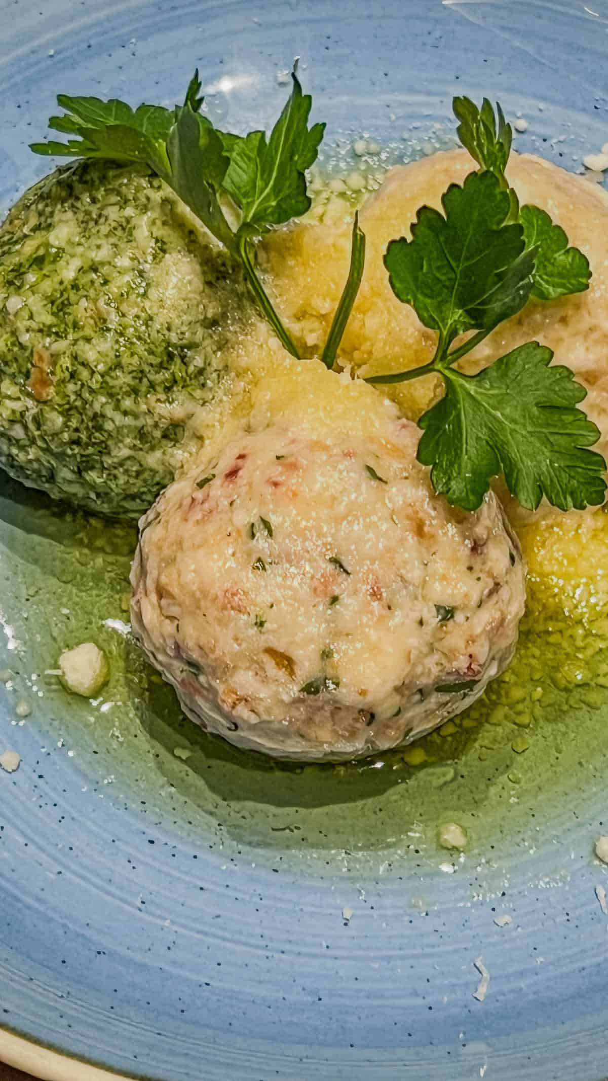 Three herb dumplings on a blue plate, garnished with parsley and set in a light buttery sauce, evoke the cozy warmth of Bolzano's Christmas Market.