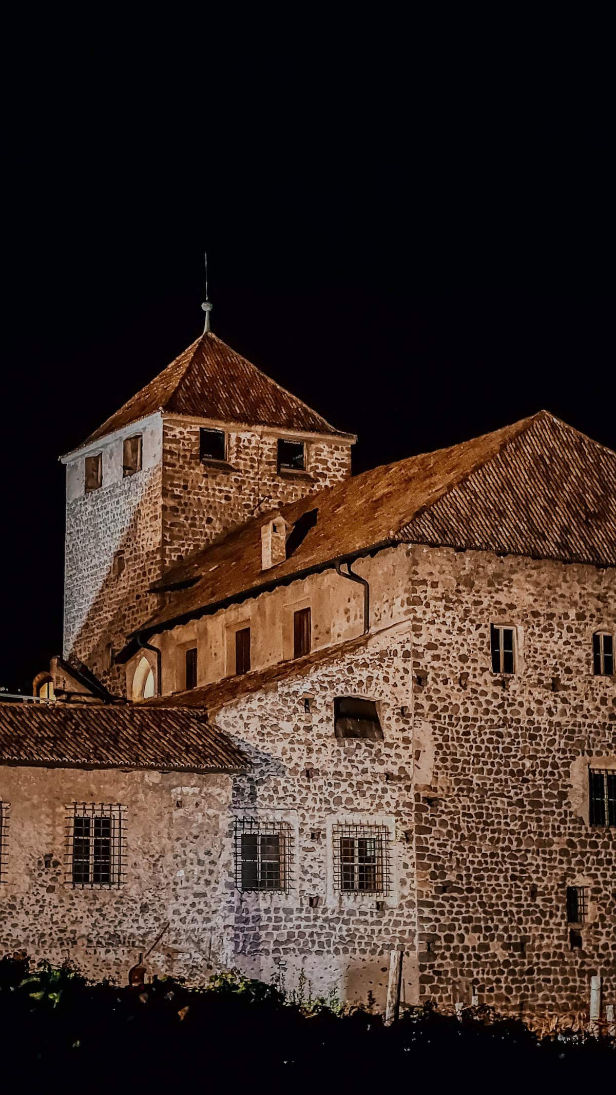 Night view of a stone building with a tower, featuring a triangular roof and small windows, reminiscent of Bolzano's charm during Christmas, set against a dark sky.