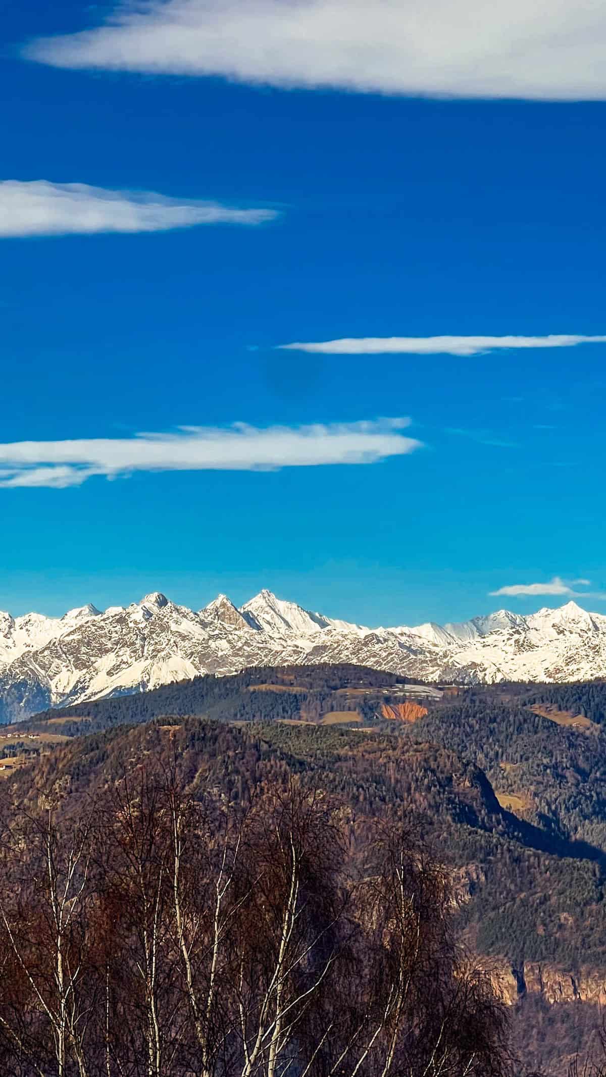 Snow-capped mountains under a clear blue sky with scattered clouds, trees in the foreground, reminiscent of the scenic beauty surrounding Bolzano during its enchanting Christmas Market season.