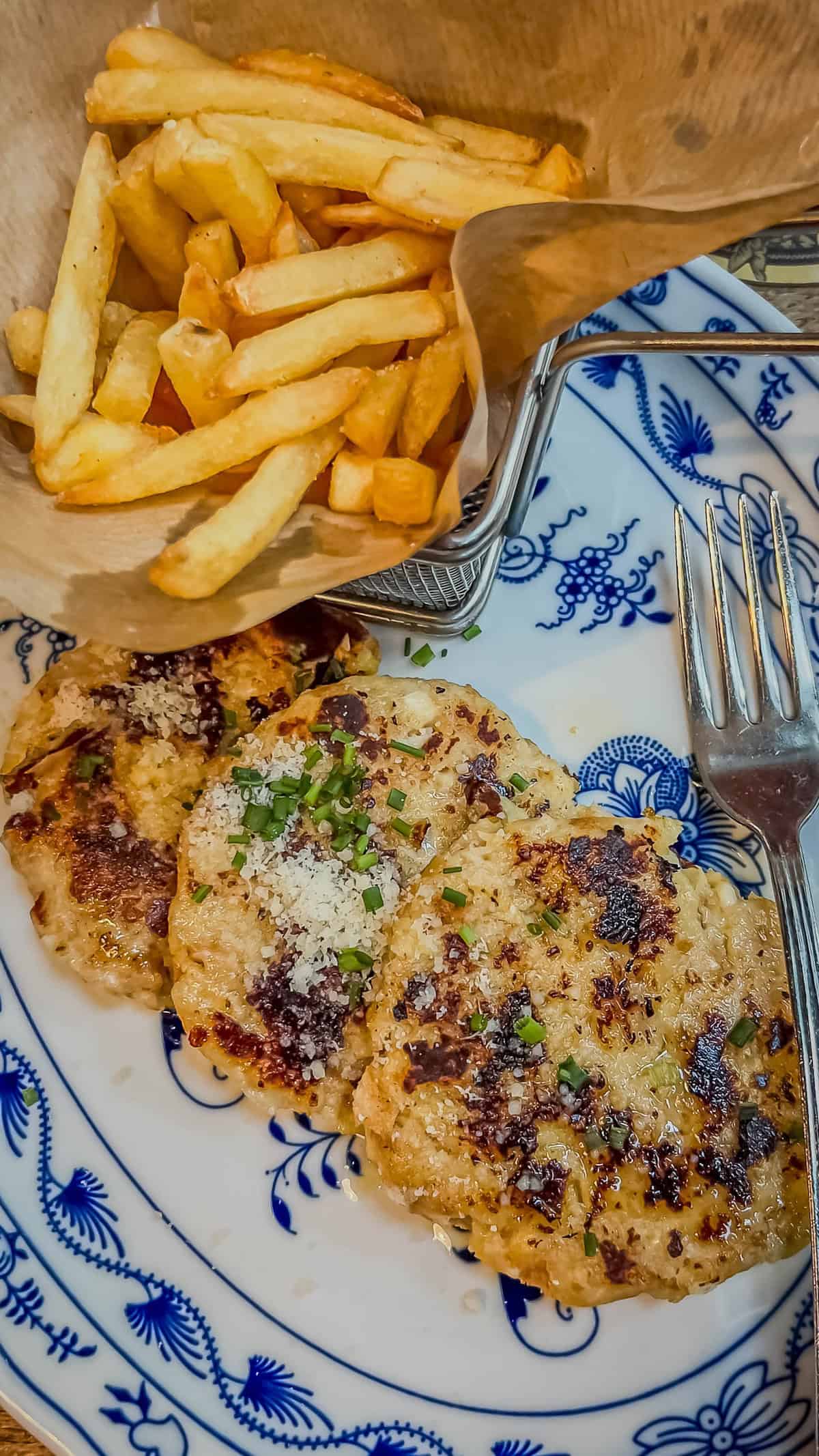 A plate with three seasoned patties garnished with herbs and Parmesan, accompanied by a fork, captures the essence of Bolzano's culinary delights. A side of golden French fries in a paper cone adds charm reminiscent of a Christmas market feast.