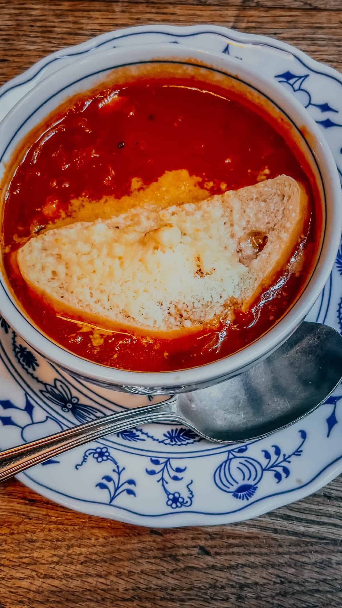 A bowl of tomato soup with a slice of bread and melted cheese on top, reminiscent of the cozy flavors found at a Christmas Market in Bolzano, served on a floral-patterned plate with a spoon beside it.