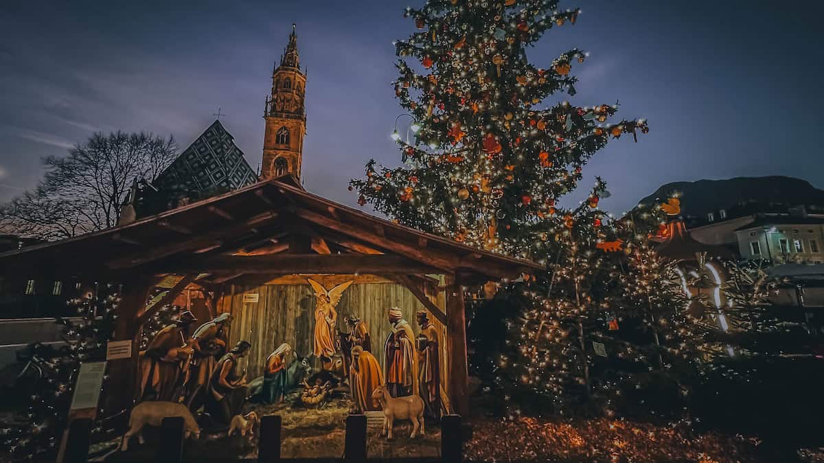 At night, the nativity scene glows beside a lit Christmas tree, with Bolzano's tall tower forming a picturesque backdrop, capturing the festive spirit of the town known for its charming Christmas Market.