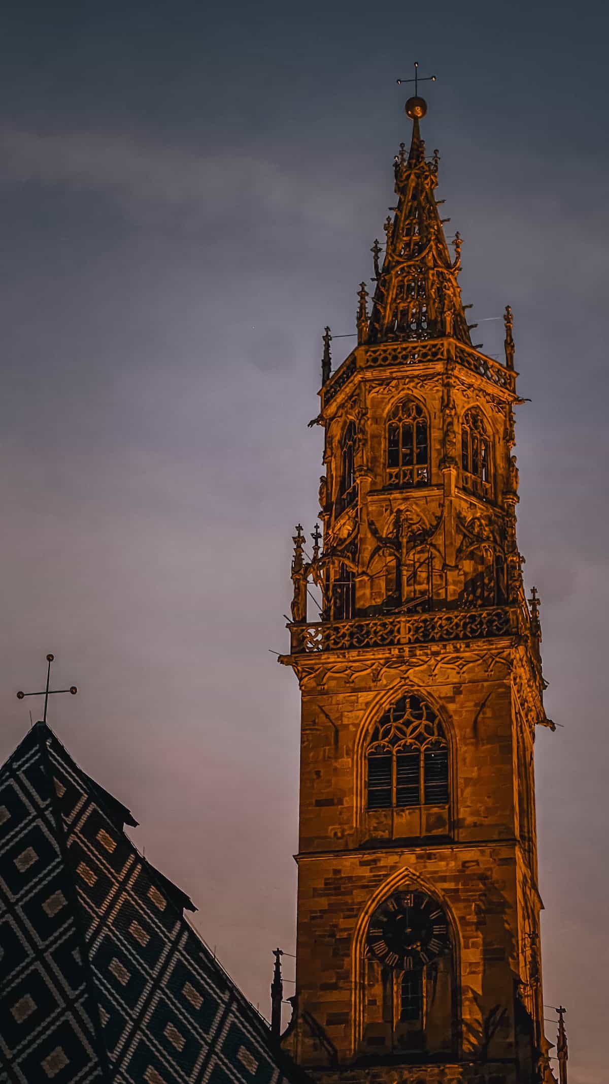 Against a dusky sky, the tall gothic-style church tower in Bolzano stands with detailed stone carvings and a clock, overlooking the festive Christmas market below.