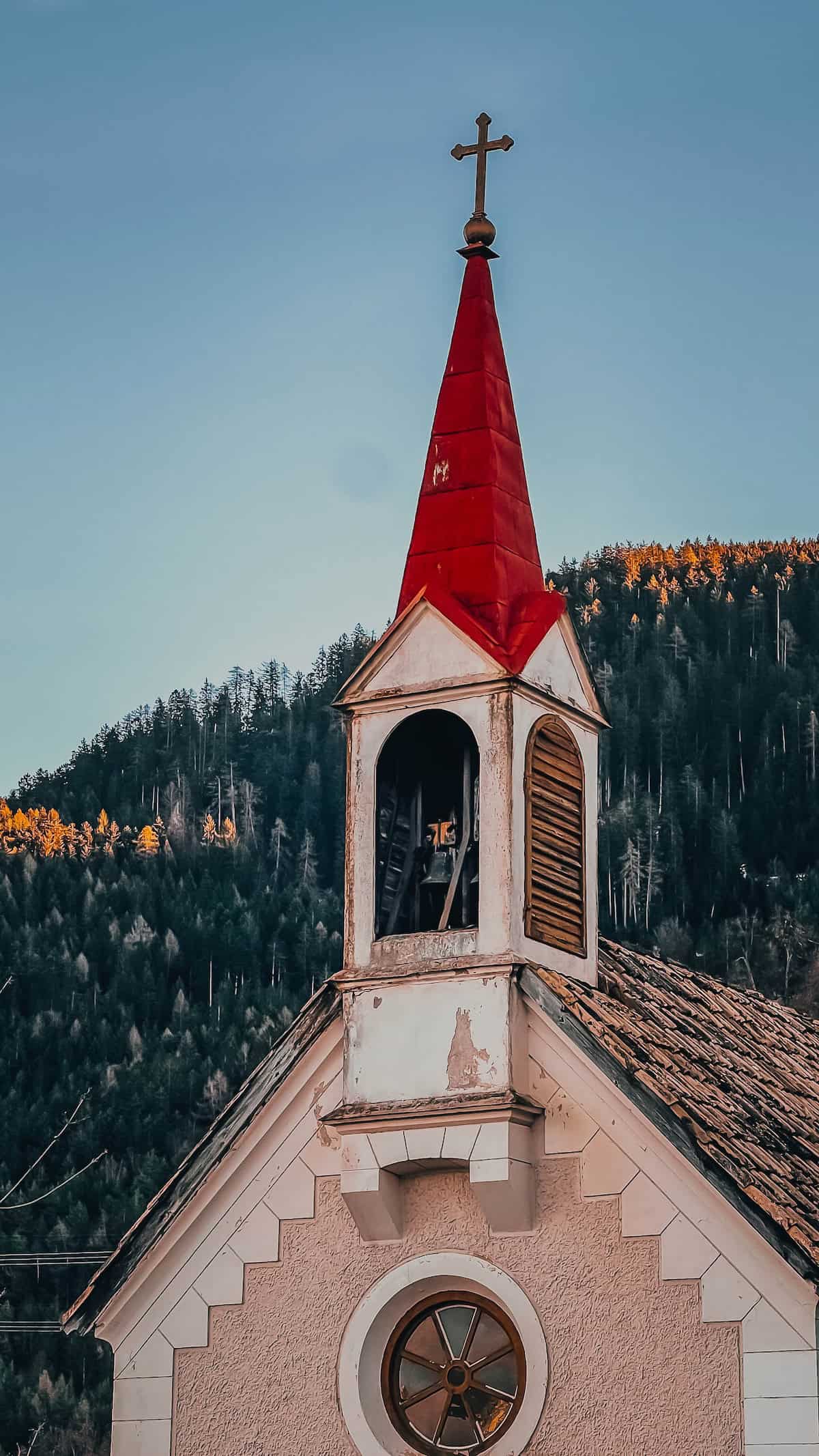 Nestled near Bolzano, this small church with a red steeple and cross stands serenely against forested hills under a clear sky, evoking the charm of a Christmas market scene.