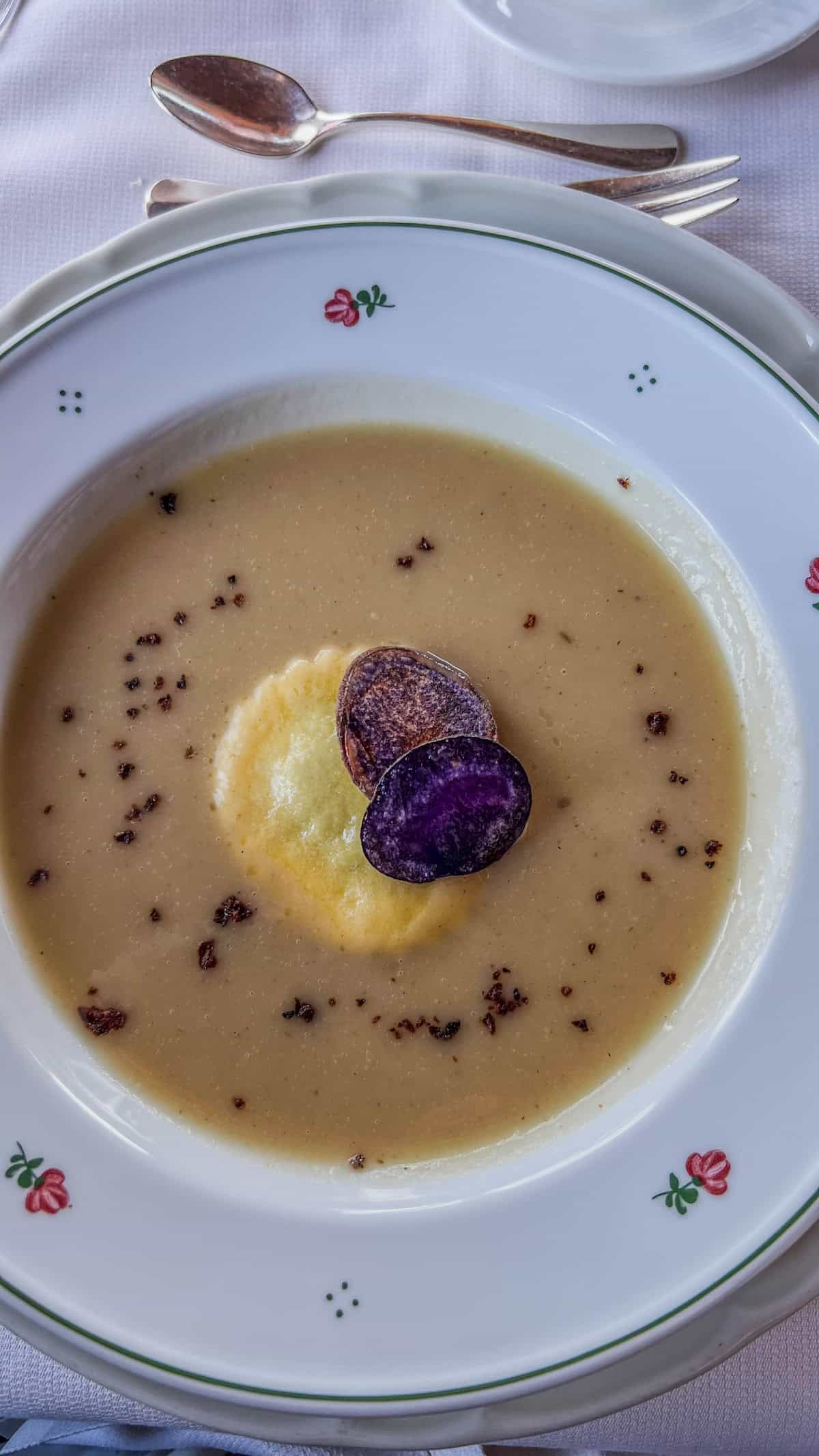 A bowl of creamy soup topped with two purple potato slices and a yellow dollop sits on a decorative white dish with a floral pattern, reminiscent of the elegance found at Bolzano's Christmas Market. Silverware is placed beside the plate.