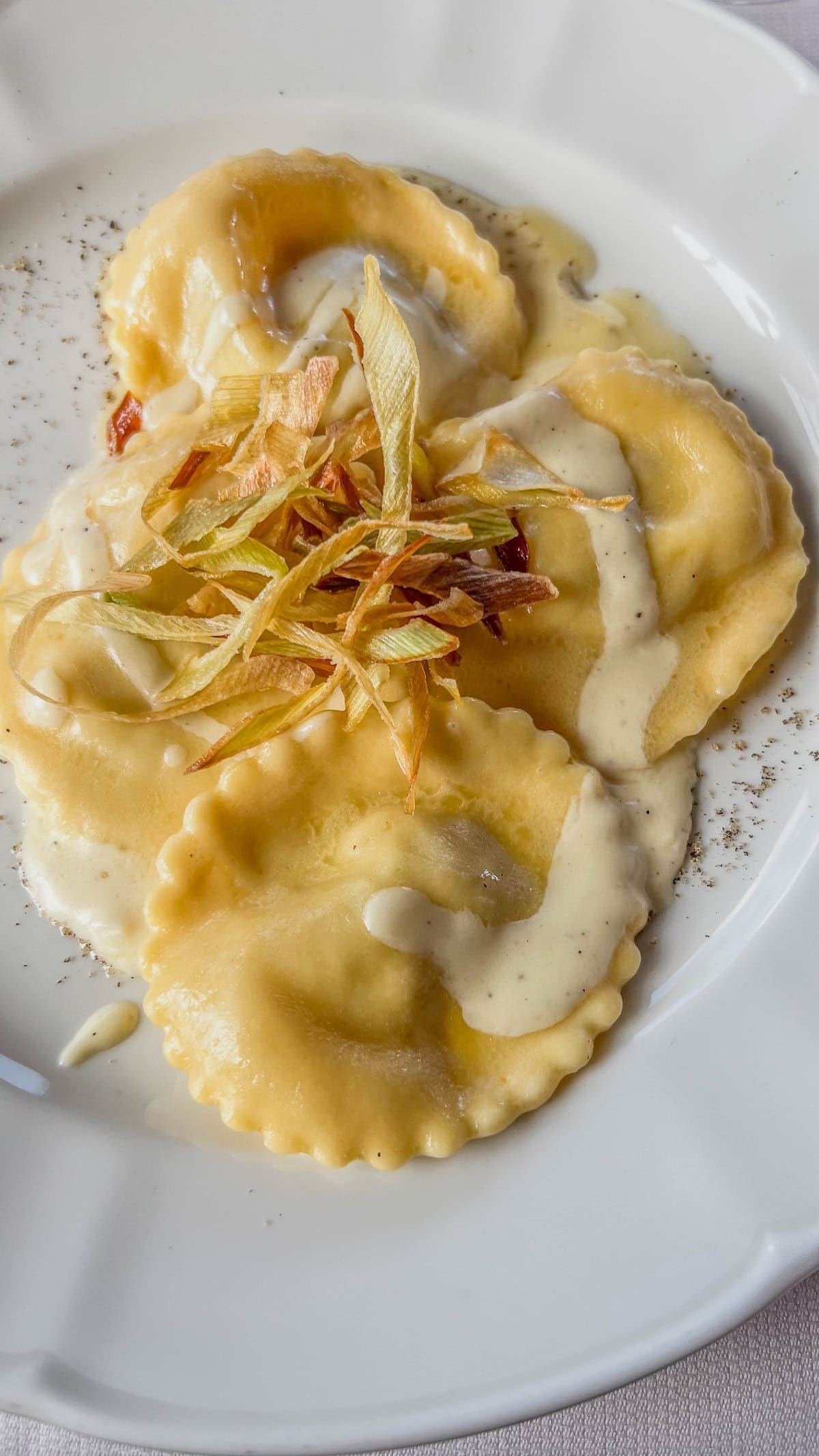 A plate of ravioli topped with creamy sauce and crispy fried onions, reminiscent of a festive Market treat in Bolzano, rests invitingly on a white dish.