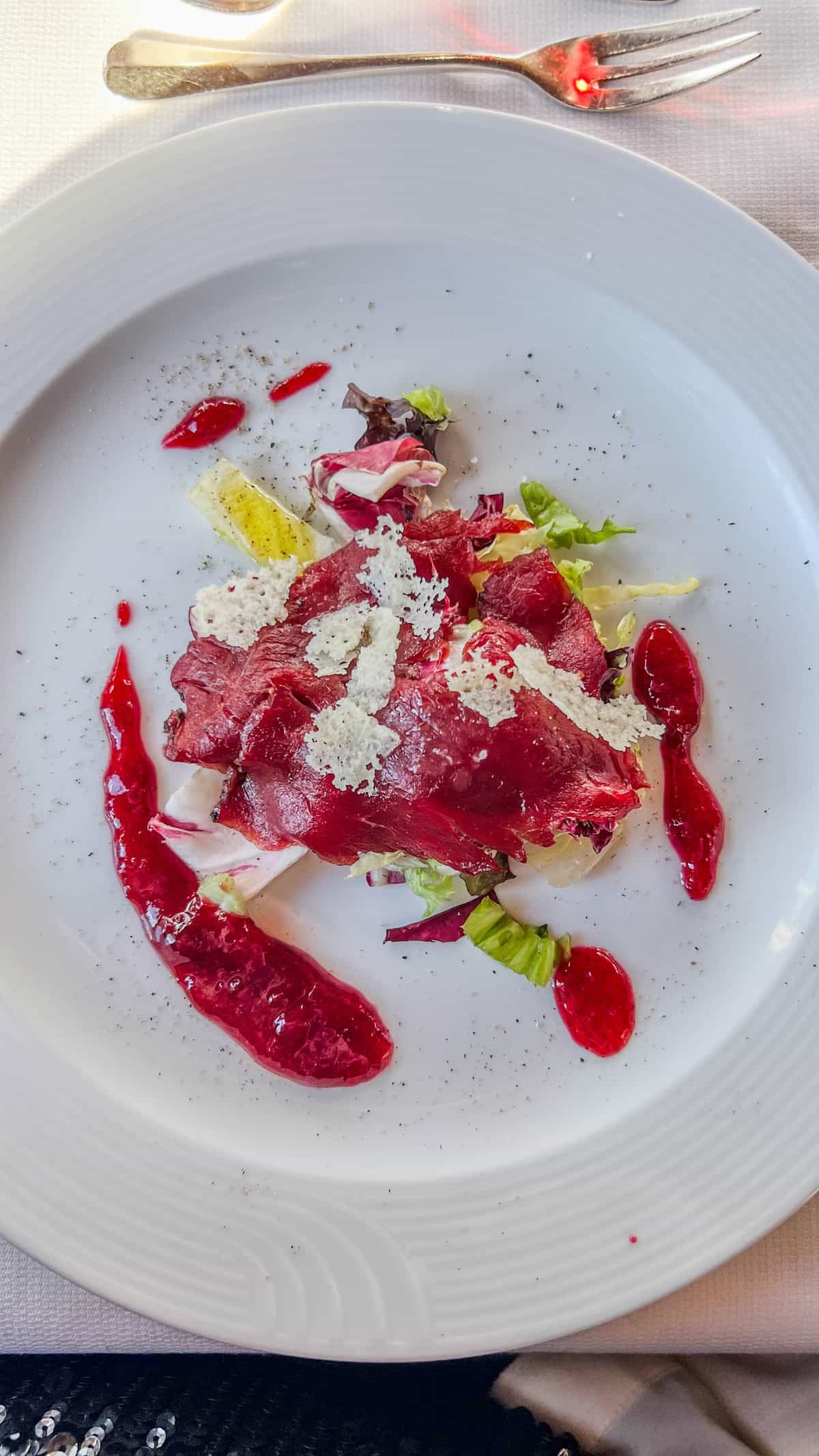 A plate of sliced beetroot with green lettuce, white cheese shavings, and red sauce drizzled on the side evokes the festive spirit of a Christmas Market in Bolzano. The vibrant dish is elegantly presented on a white plate, with a fork placed at the top right.