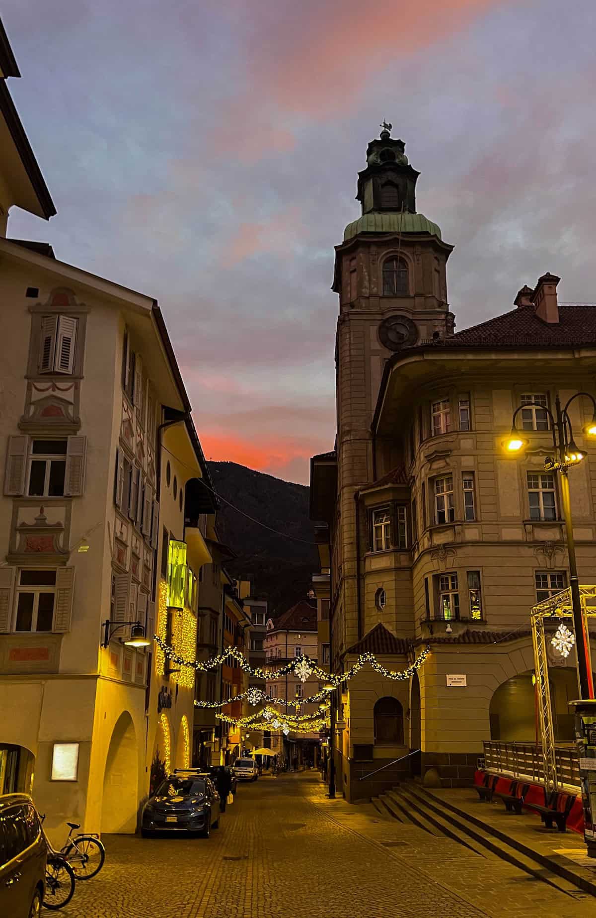 A narrow cobblestone street lined with buildings and festive lights, reminiscent of a cozy Christmas market in Bolzano, features a clock tower set against a colorful sunset sky.