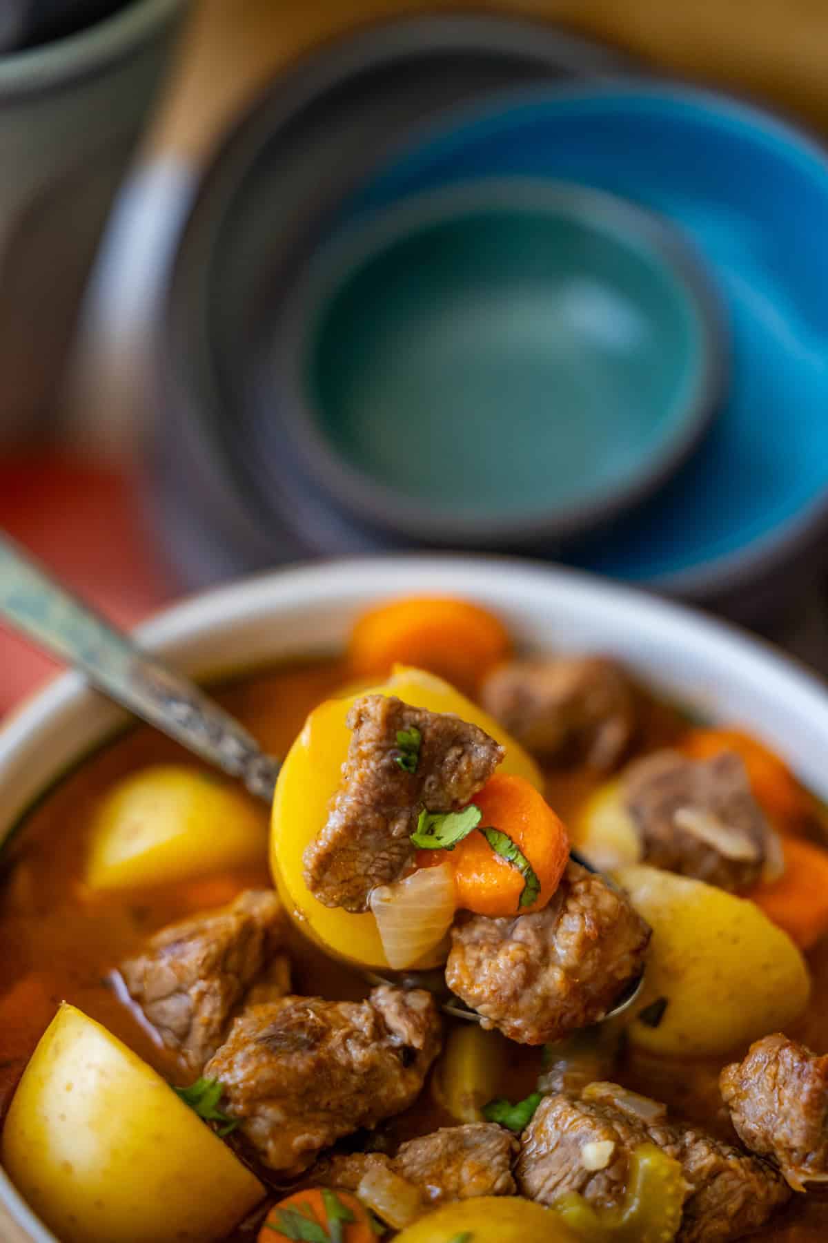 Close-up of a bowl of classic beef stew containing chunks of beef, potatoes, carrots, and onions, garnished with fresh herbs, with a spoon placed in the bowl. Stacked bowls are visible in the background.