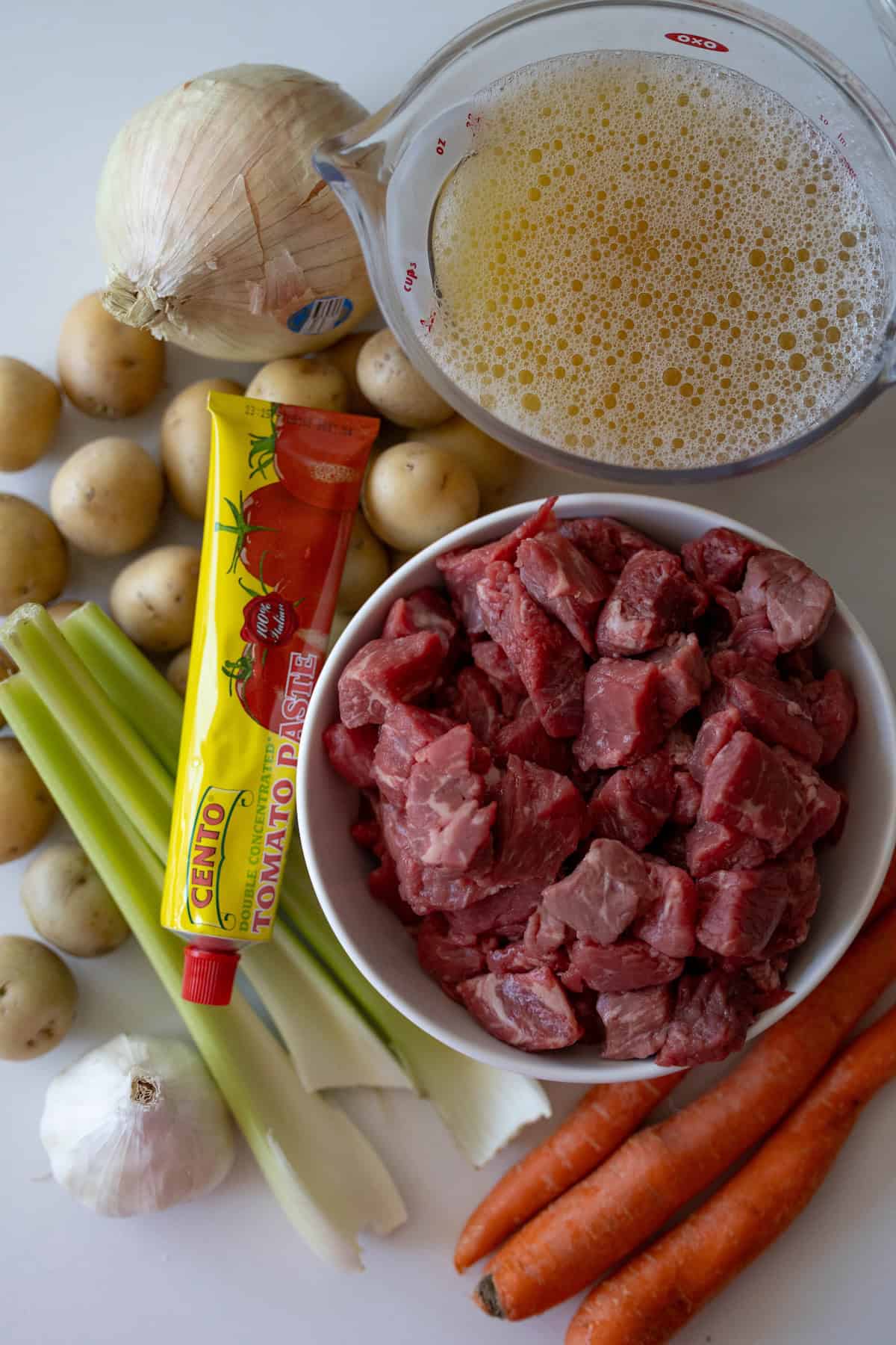 Ingredients for a classic beef stew include raw beef chunks in a white bowl, a tube of tomato paste, celery stalks, an onion, carrots, garlic, small potatoes, and a measuring cup filled with broth.