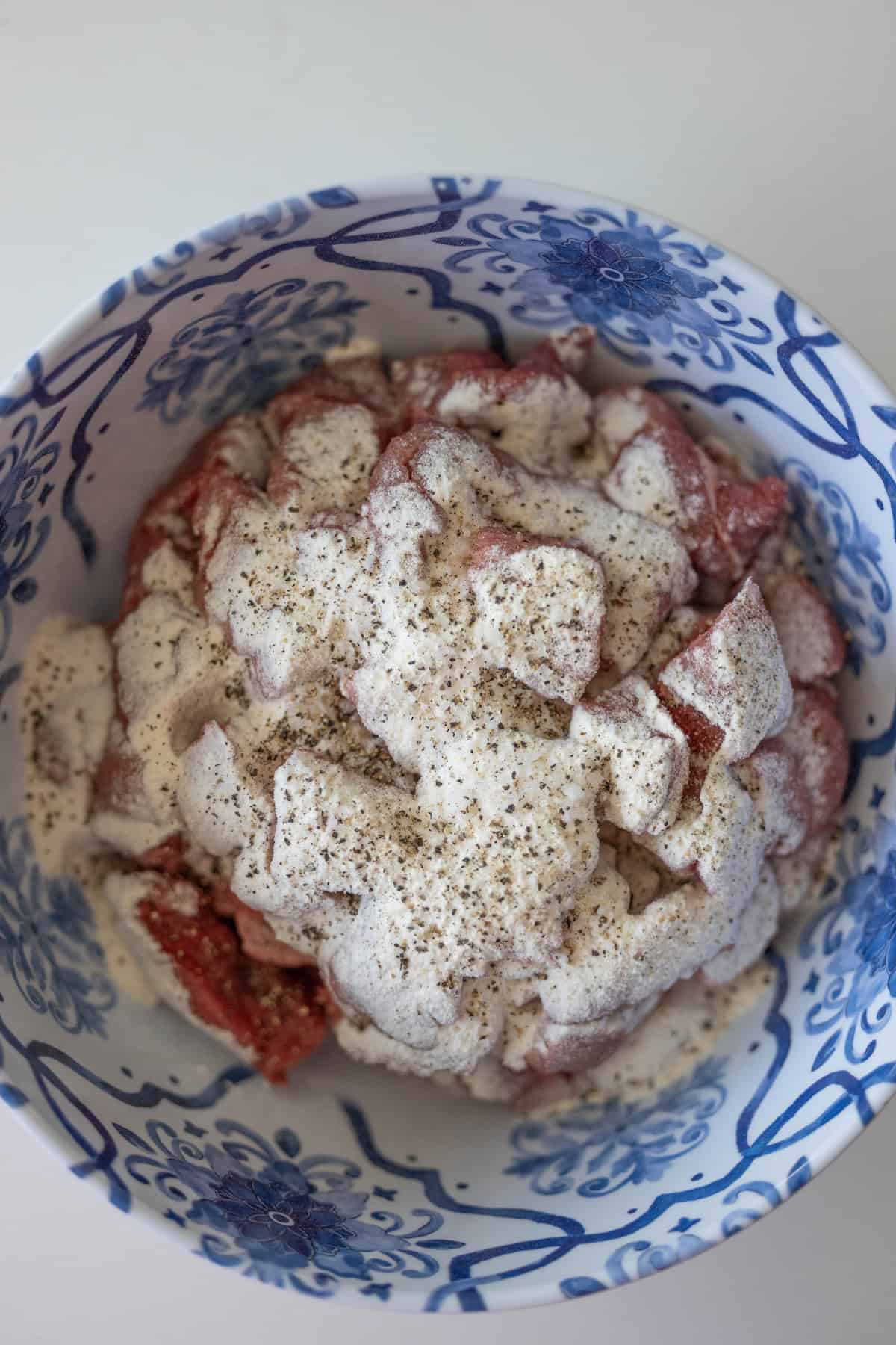 A bowl containing raw meat pieces covered with a white creamy marinade and sprinkled with black pepper suggests the beginning of a classic beef stew. The bowl, decorated with blue floral patterns, adds an elegant touch.