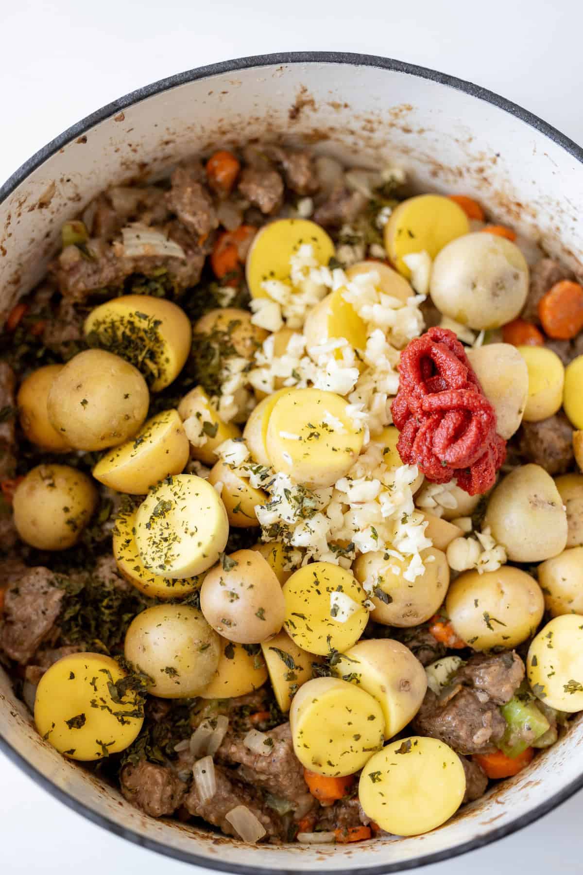 A pot contains yellow baby potatoes, minced garlic, tomato paste, diced carrots, and meat, all seasoned with green herbs for a classic beef stew.