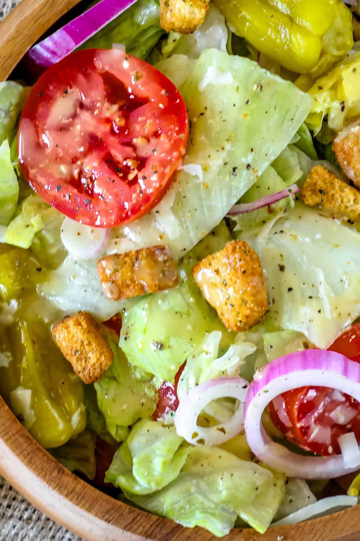 A close-up image of a fresh salad featuring sliced tomatoes, onion rings, croutons, chopped lettuce, and pepperoncini, in a wooden bowl.