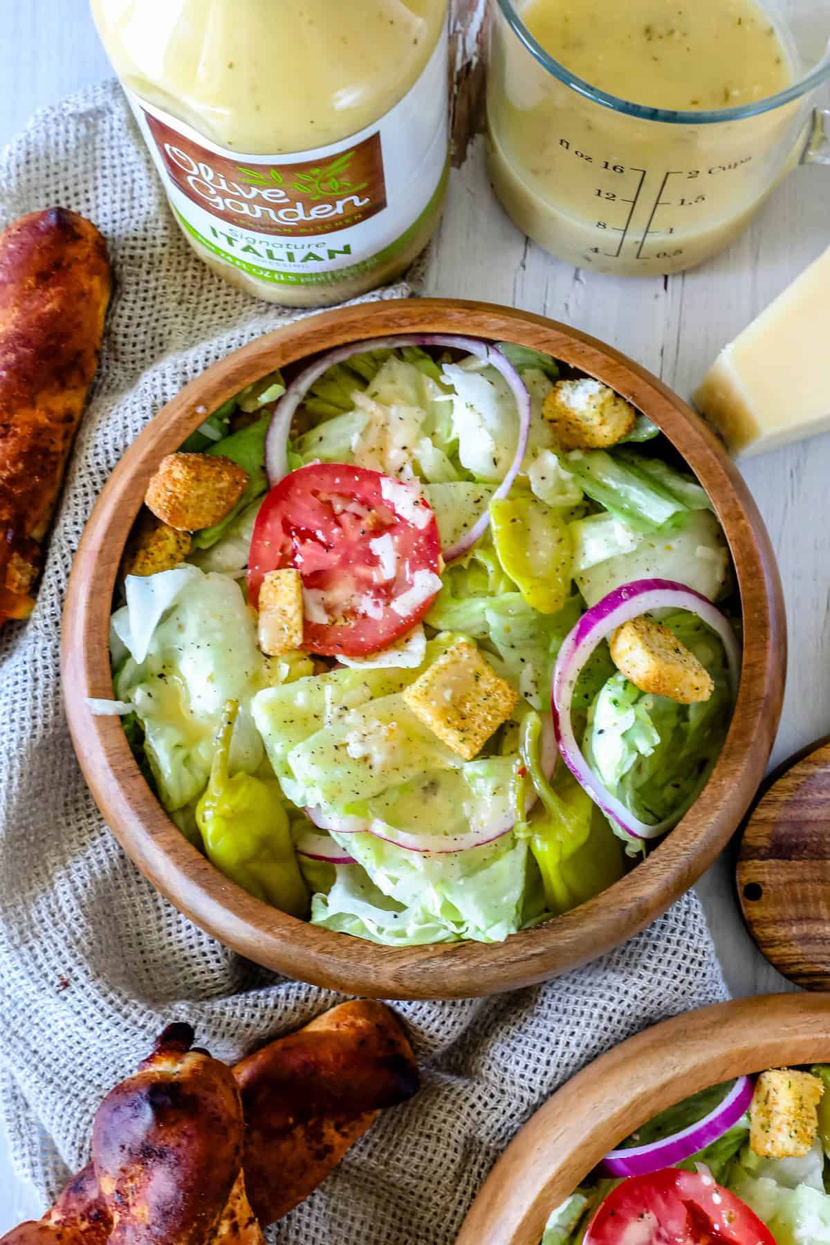 A wooden bowl filled with a salad containing lettuce, tomato slices, red onion, croutons, and grated cheese, placed beside breadsticks, a bottle of Olive Garden dressing, and a measuring cup with dressing.