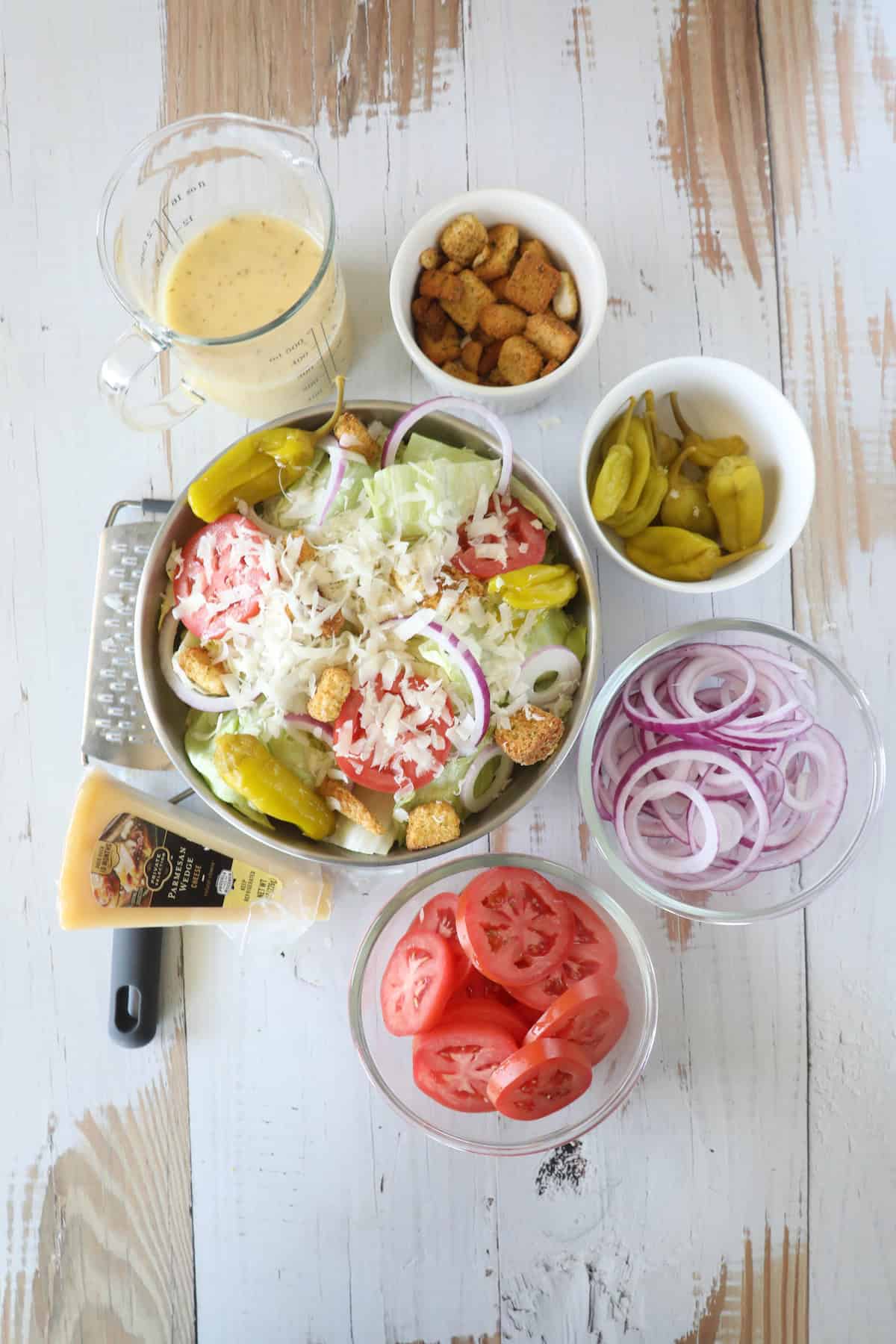 A salad with shredded cheese, pepperoncini, tomatoes, onions, and croutons in a bowl. Surrounding the bowl are separate dishes of tomatoes, onion slices, croutons, pepperoncini, cheese, and dressing.