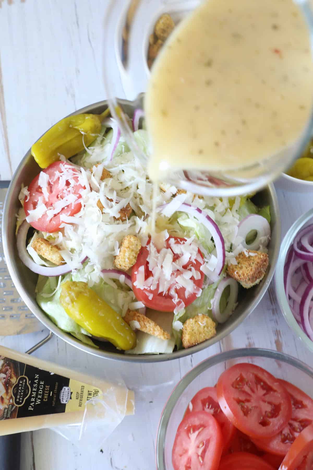 A bowl of salad with lettuce, tomatoes, onions, pepperoncini, shredded cheese, and croutons. Salad dressing is being poured over it. Parmesan cheese and bowls of sliced tomatoes and onions are nearby.