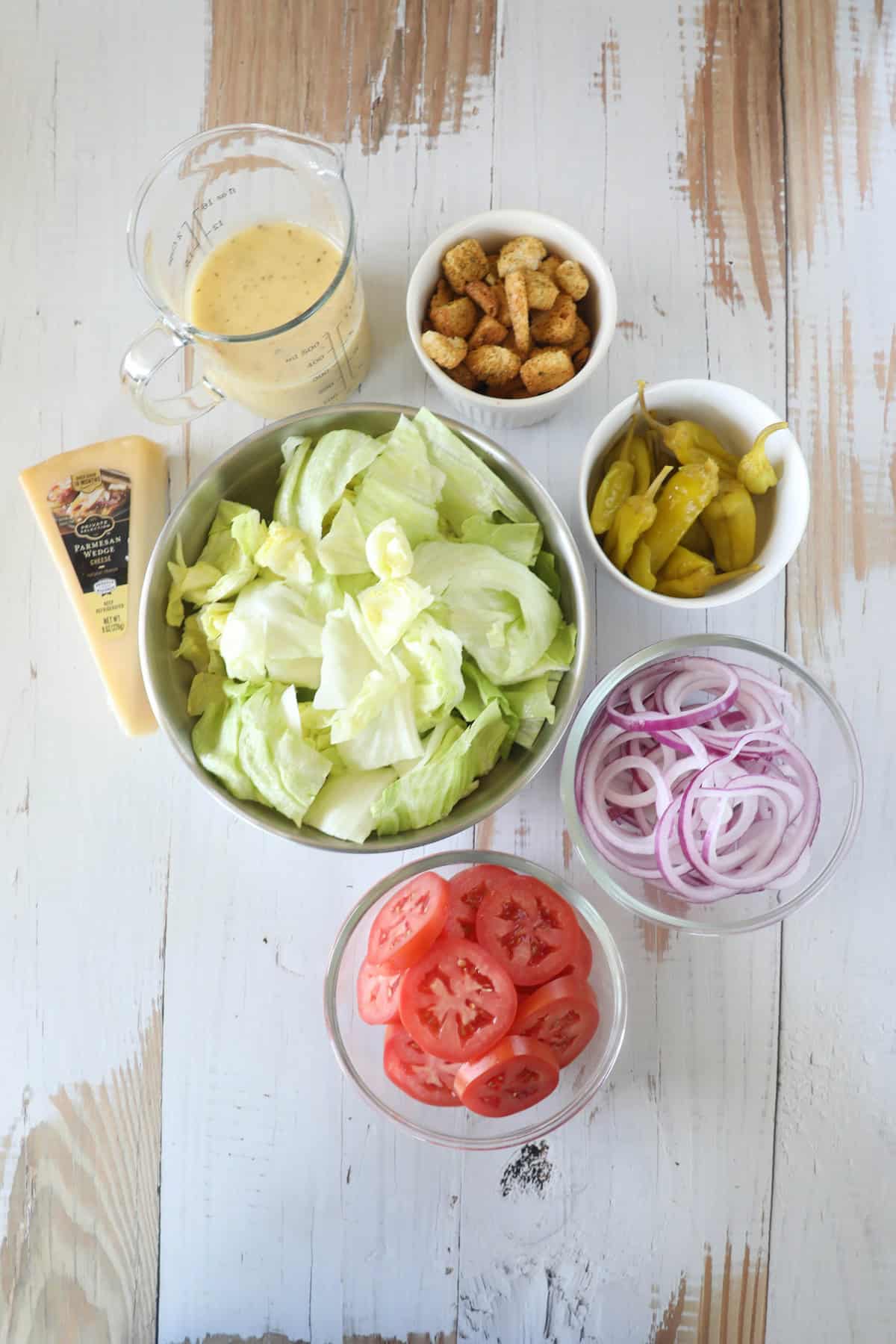 Ingredients for a salad including a bowl of lettuce, sliced tomatoes, sliced red onions, peppers, croutons, a small jug of dressing, and a tube of Caesar salad dressing on a wooden surface.