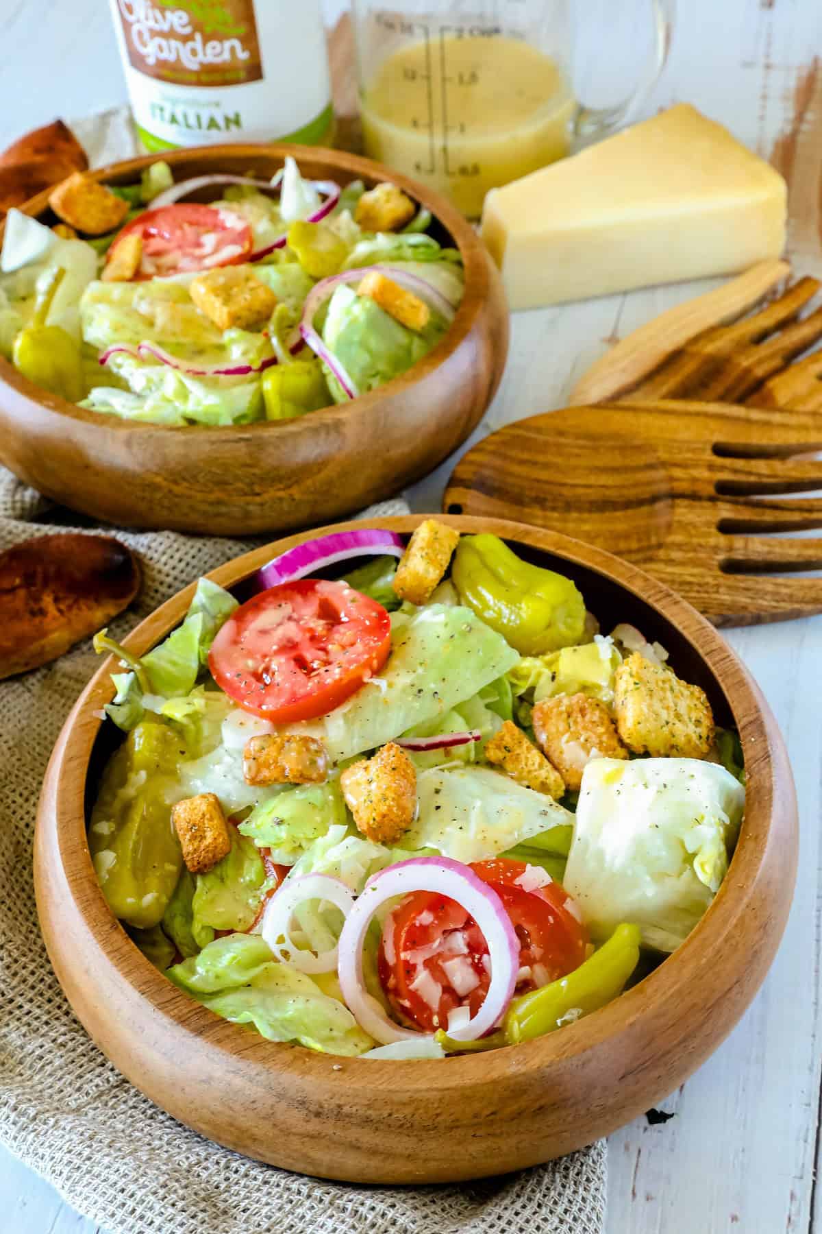 Two wooden bowls filled with fresh salad featuring lettuce, tomatoes, red onions, pepperoncini, and croutons, with dressing, cheese, and salad utensils in the background.