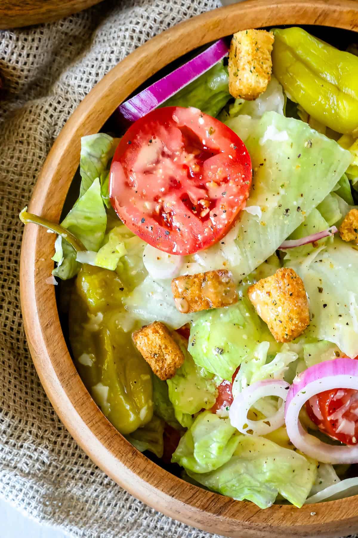 A wooden bowl filled with a salad consisting of lettuce, sliced tomato, croutons, pepperoncini, and red onion, all drizzled with a light dressing.