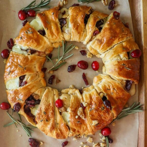 A golden-brown cranberry brie wreath features a pastry ring filled with cheese and cranberries, garnished with rosemary sprigs and fresh cranberries atop parchment paper.