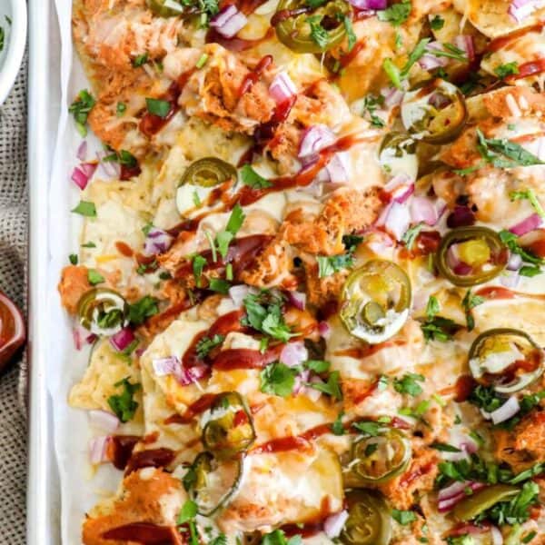 A tray of BBQ Chicken Nachos topped with shredded cheese, barbecue sauce, red onions, jalapeños, and cilantro. Try this mouthwatering recipe for your next gathering!