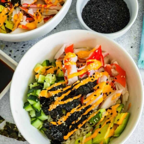 A tempting bowl of sushi salad featuring rice, cucumber, avocado, crab sticks, seaweed strips, and drizzled with orange sauce—reminiscent of California Roll Bowls. A small bowl of black sesame seeds is visible in the background, perfect for adding an extra touch to this delectable recipe.