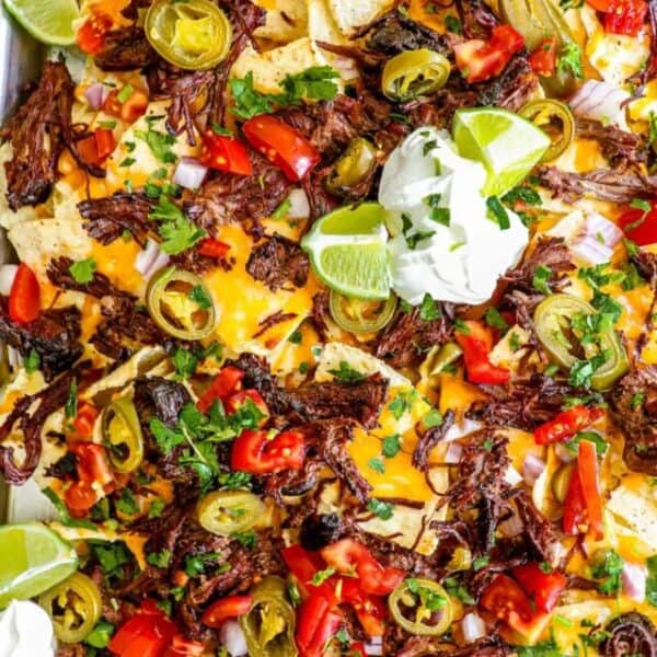 A tray of loaded nachos topped with juicy carne asada, melted cheese, sliced jalapeños, tomatoes, diced onions, cilantro, lime wedges, and dollops of sour cream offers a perfect twist on a classic recipe.