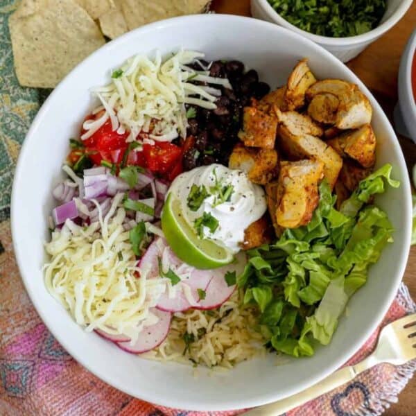 A Chicken Burrito Bowl that’s picture-perfect: a bowl of rice topped with grilled chicken, shredded cheese, black beans, chopped tomatoes, sliced radishes, lettuce, chopped onions, cilantro, sour cream, and a lime wedge. Tortilla chips and cilantro on the side complete this delicious recipe.