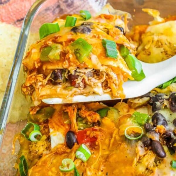 A serving of chicken enchilada is being lifted from a glass baking dish with a white spatula. The enchilada is covered in melted cheese, diced green onions, black beans, and green bell peppers.