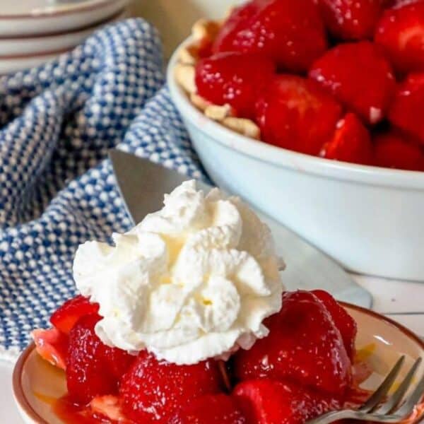 A plate of strawberry pie topped with whipped cream is in the foreground, showcasing an easy recipe, with a large bowl of strawberries and a container labeled "sugar" in the background.