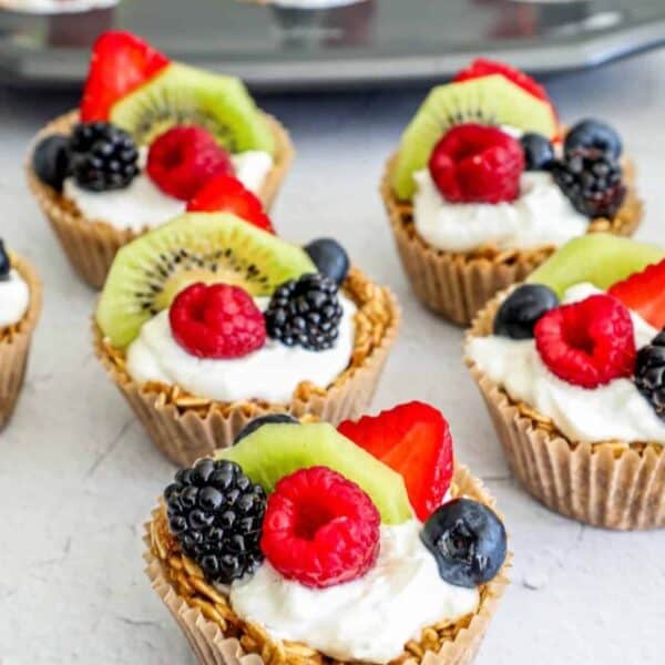 Mini fruit tartlets with a granola crust, topped with creamy yogurt and an assortment of fresh raspberries, strawberries, blueberries, blackberries, and kiwi slices. A delightful recipe showcasing additional tartlets in the background in a muffin tin.