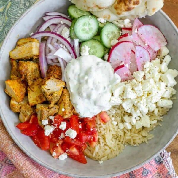 A picture-perfect Greek Chicken Bowl featuring seasoned chicken cubes, sliced red onions, cucumber, radishes, tomatoes, crumbled feta, rice, and a dollop of tzatziki. A piece of naan bread graces the edge of the bowl—a true recipe for delight.