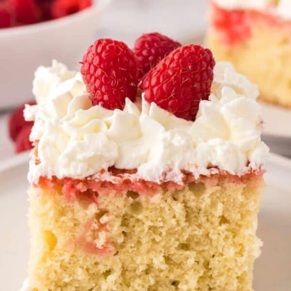 A slice of raspberry sheet cake topped with whipped cream and fresh raspberries on a white plate.