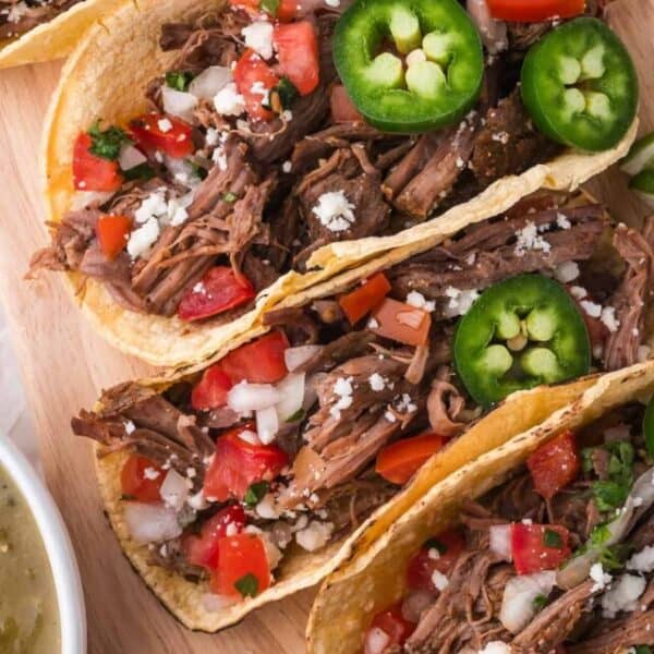 Three shredded beef tacos in corn tortillas garnished with diced tomatoes, onions, crumbled cheese, and sliced jalapeños, served on a wooden board with a side of green salsa.