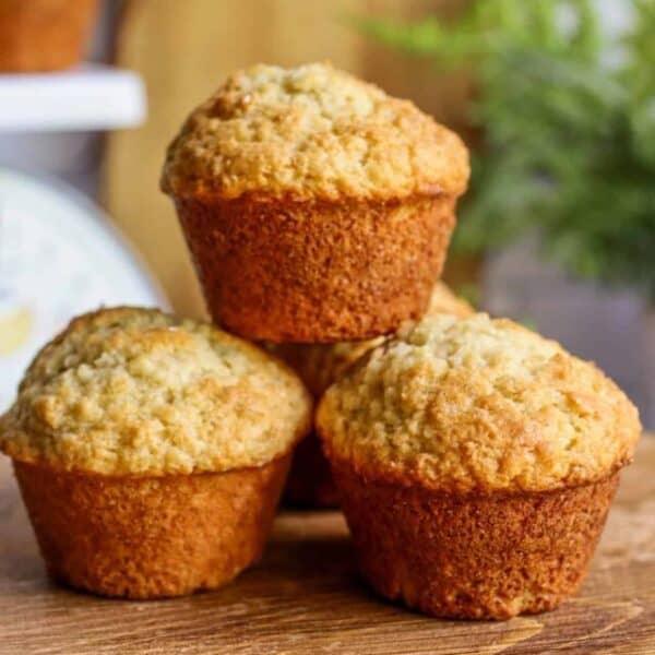 Three freshly baked sourdough muffins are stacked on a wooden surface with a blurred background.