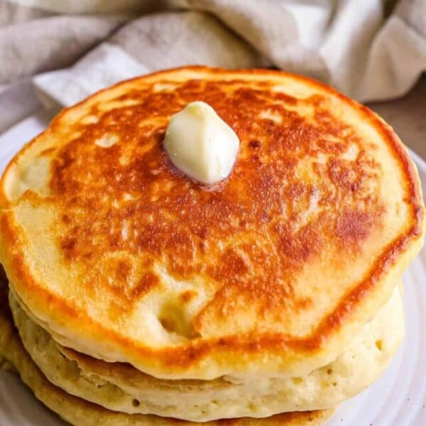 A stack of three golden-brown sourdough pancakes topped with a dollop of butter, served on a white plate, with a beige cloth in the background.