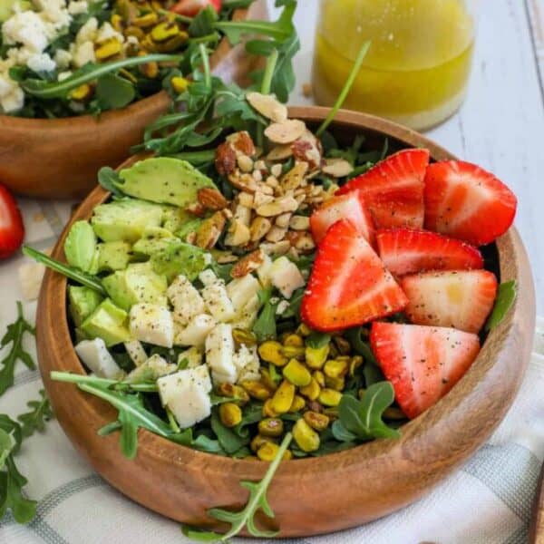 Two wooden bowls filled with strawberry crunch salads containing strawberries, avocado, nuts, cheese, and greens, accompanied by a dressing on the side.