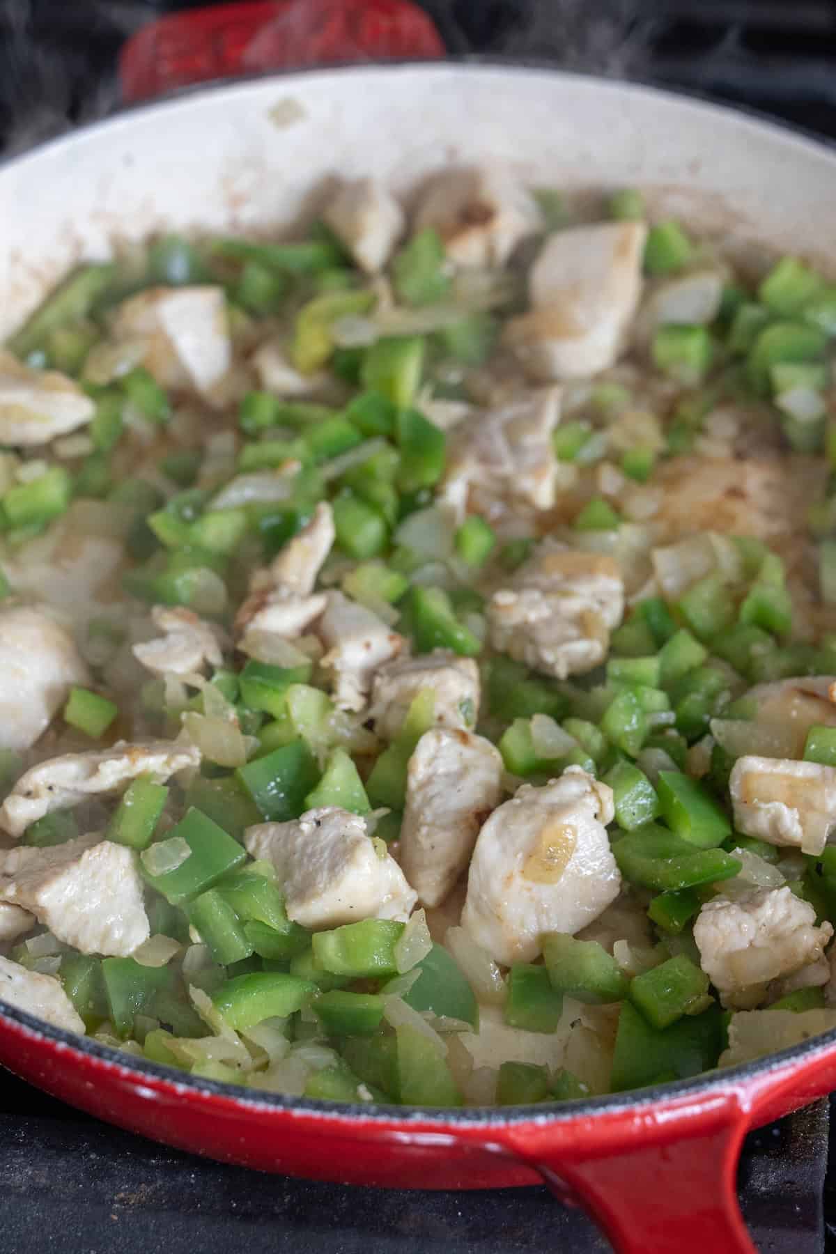 A red skillet contains a mixture of chopped green bell peppers, diced onions, and pieces of chicken cooking together, perfect for starting an easy risotto recipe.