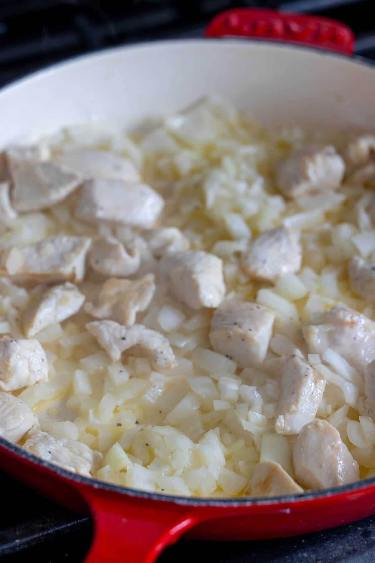 A red skillet on a stove contains pieces of cooked chicken and chopped onions, perfect for starting your one pan chicken risotto.