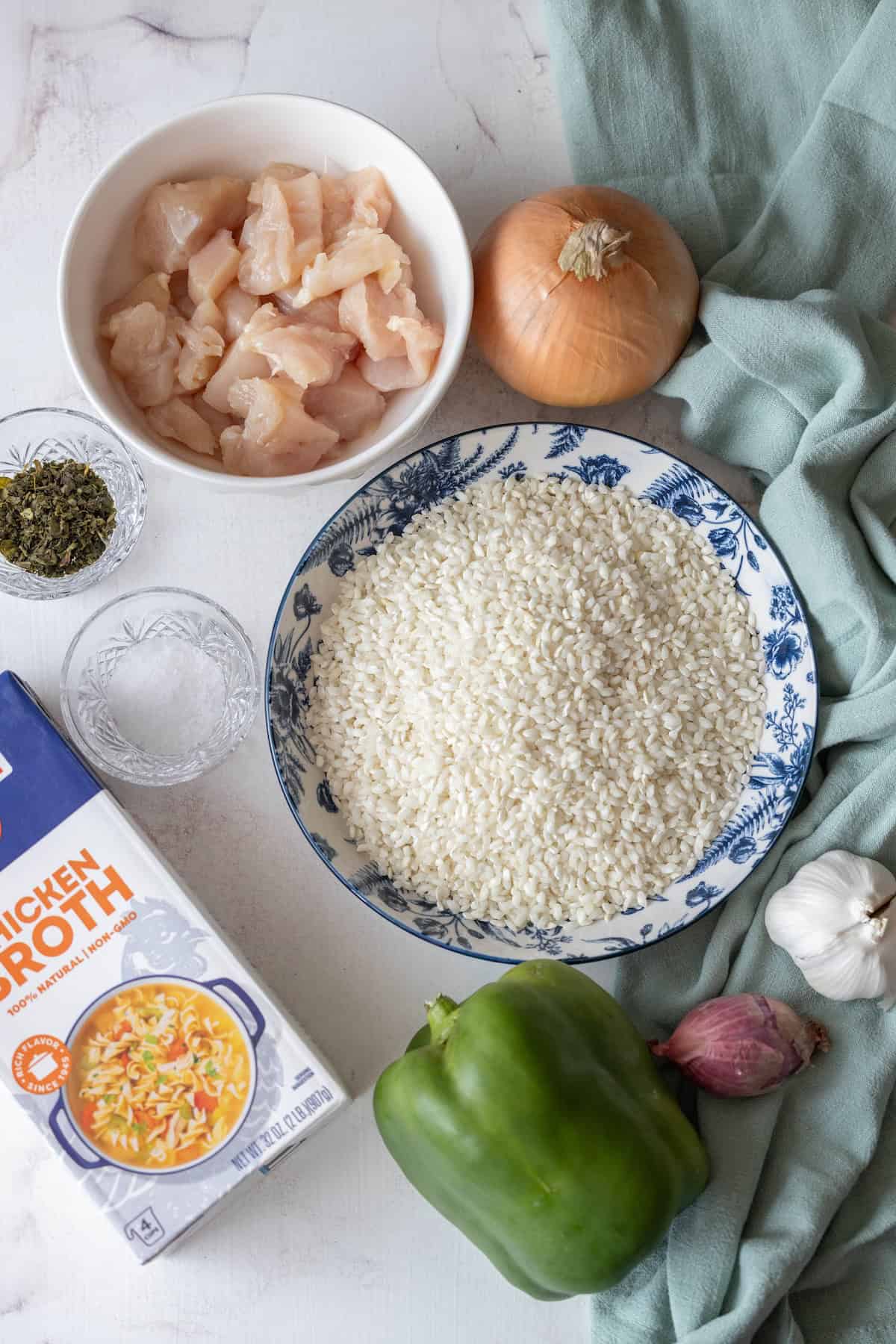 Ingredients for a chicken risotto recipe are laid out: diced chicken, white rice, chicken broth, a green bell pepper, an onion, a shallot, garlic cloves, dried herbs, and a green cloth.