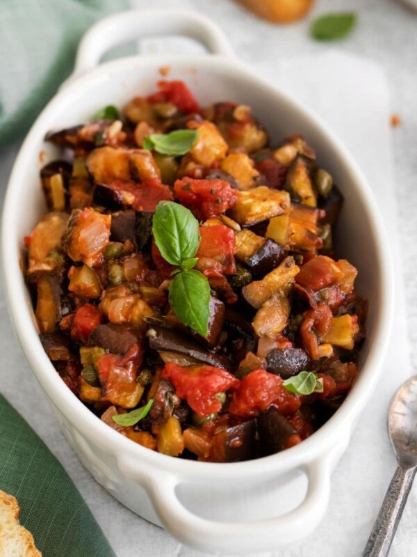 picture of baked eggplant caponata in a white dish with basil leaves on top on a table next to a spoon