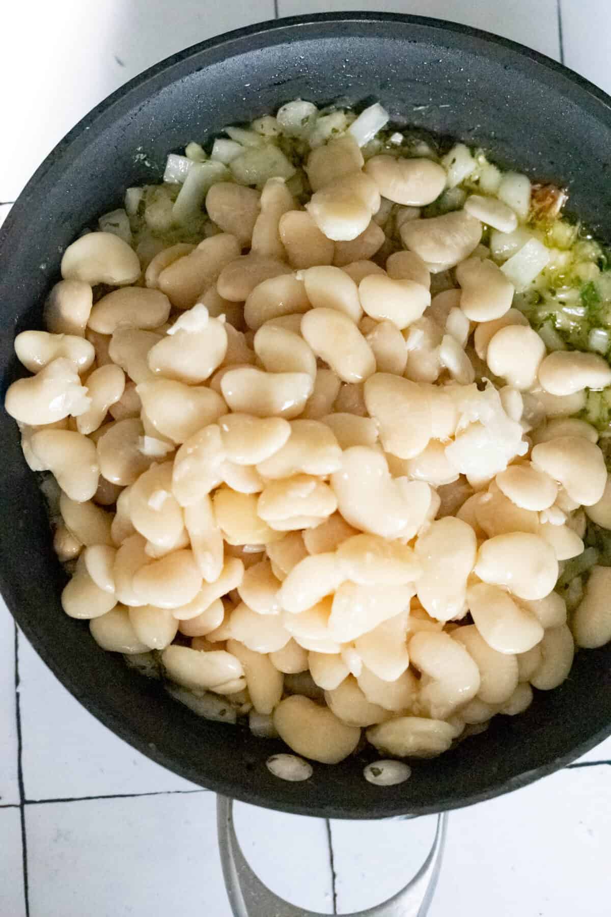 A frying pan sizzles with chopped onions, garlic, and butter beans, creating an aromatic blend.