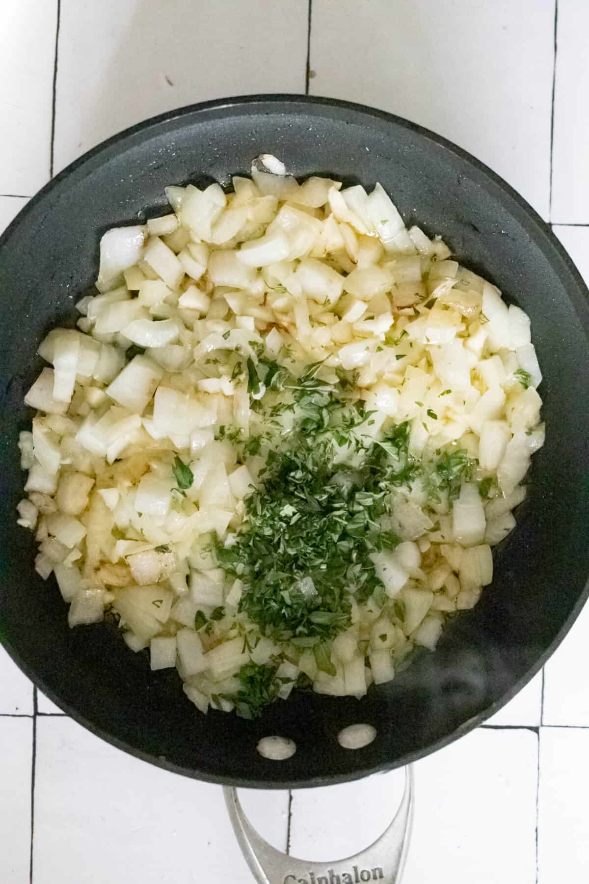 A frying pan with chopped onions, garlic, and herbs sits on a white tiled surface.